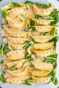 Rows of dumplings arranged in a baking dish with leafy greens and covered in a creamy orange sauce.
