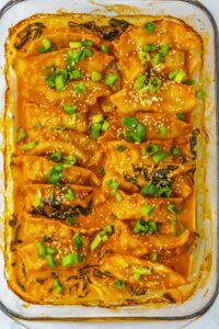 Baked dumplings in an orange sauce, topped with chopped green onions and sesame seeds, arranged in a glass baking dish.