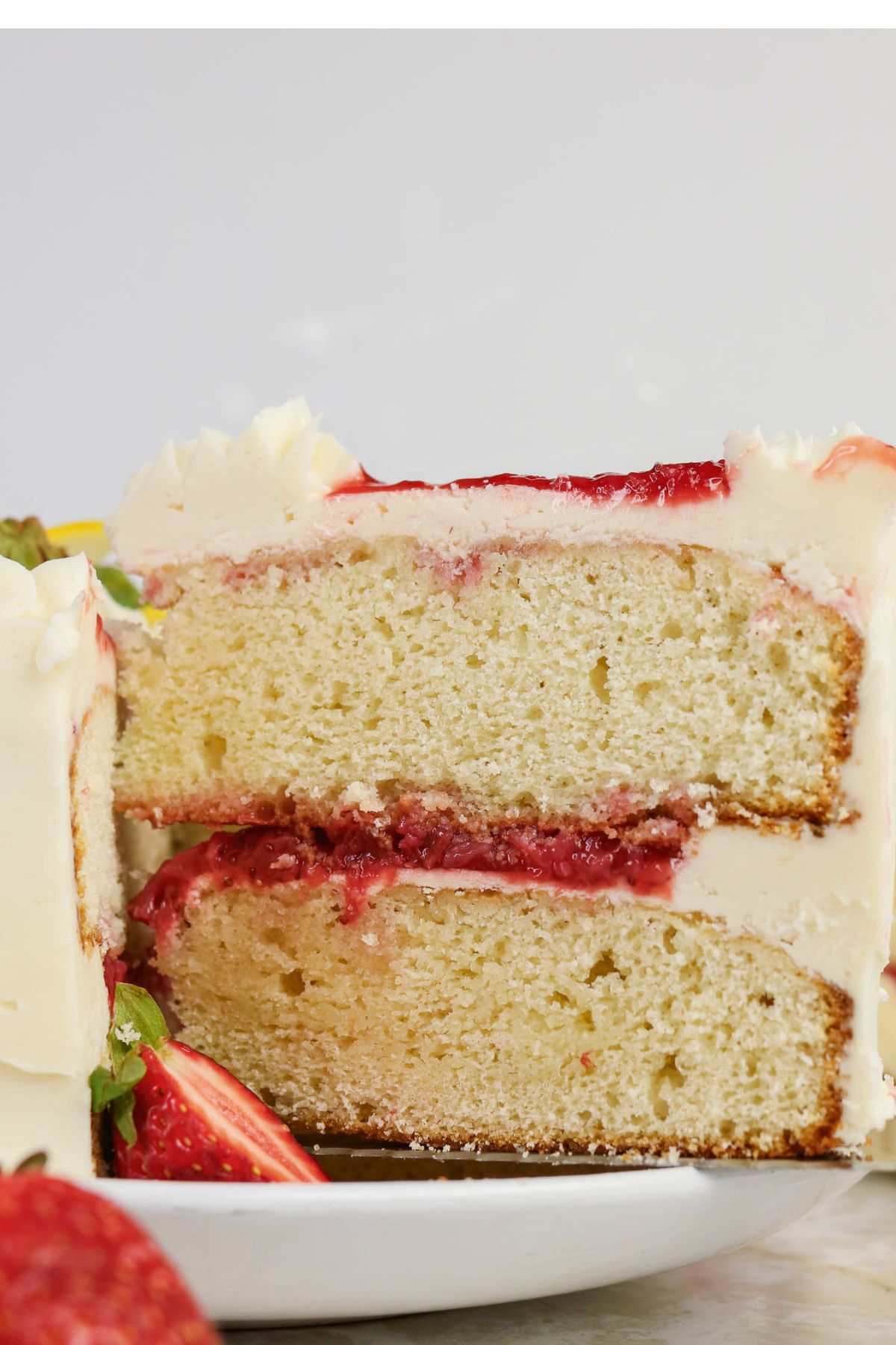 A close-up of a sliced vanilla layer cake with white frosting and a strawberry filling, displayed on a white plate with fresh strawberries.