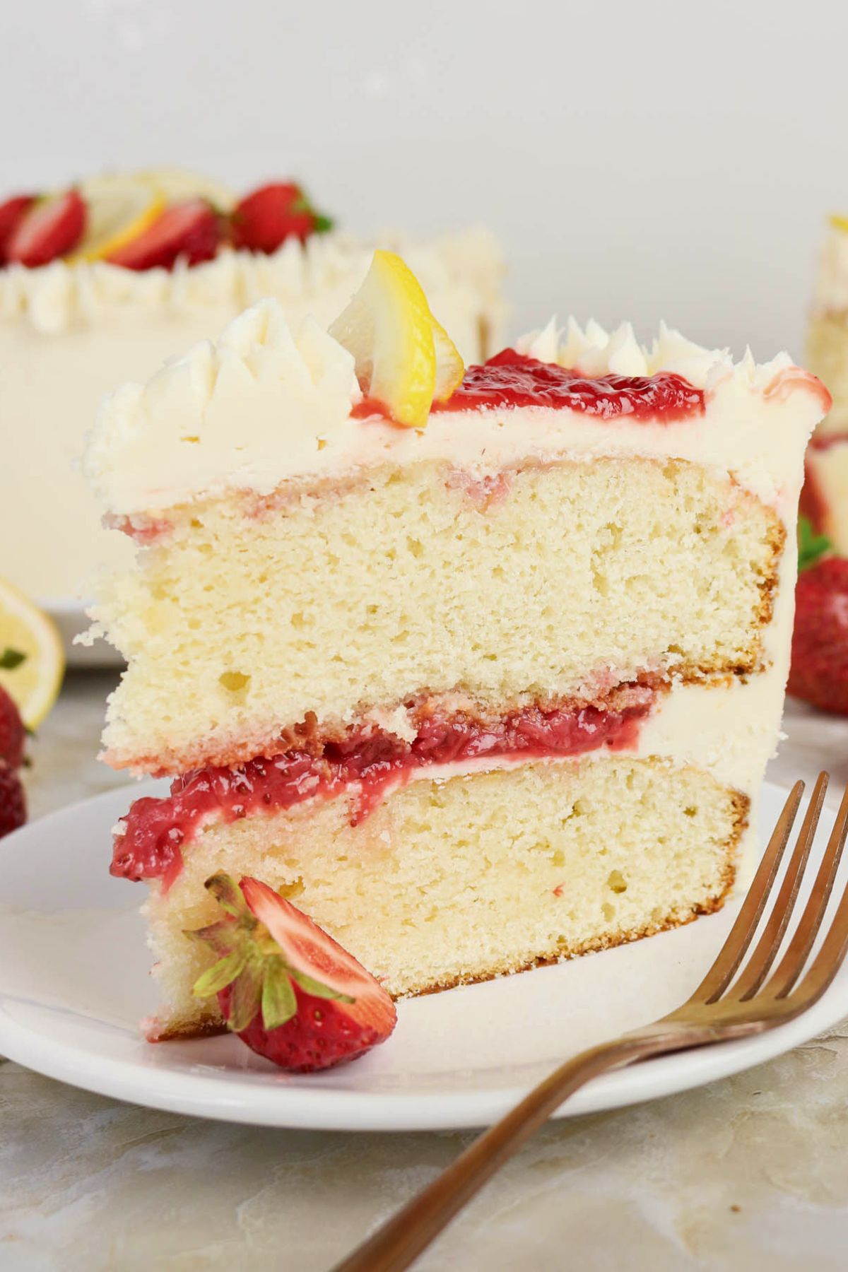 A slice of layered vanilla cake with white frosting, strawberry filling, and a lemon slice on top, served on a plate with a fork and fresh strawberries.