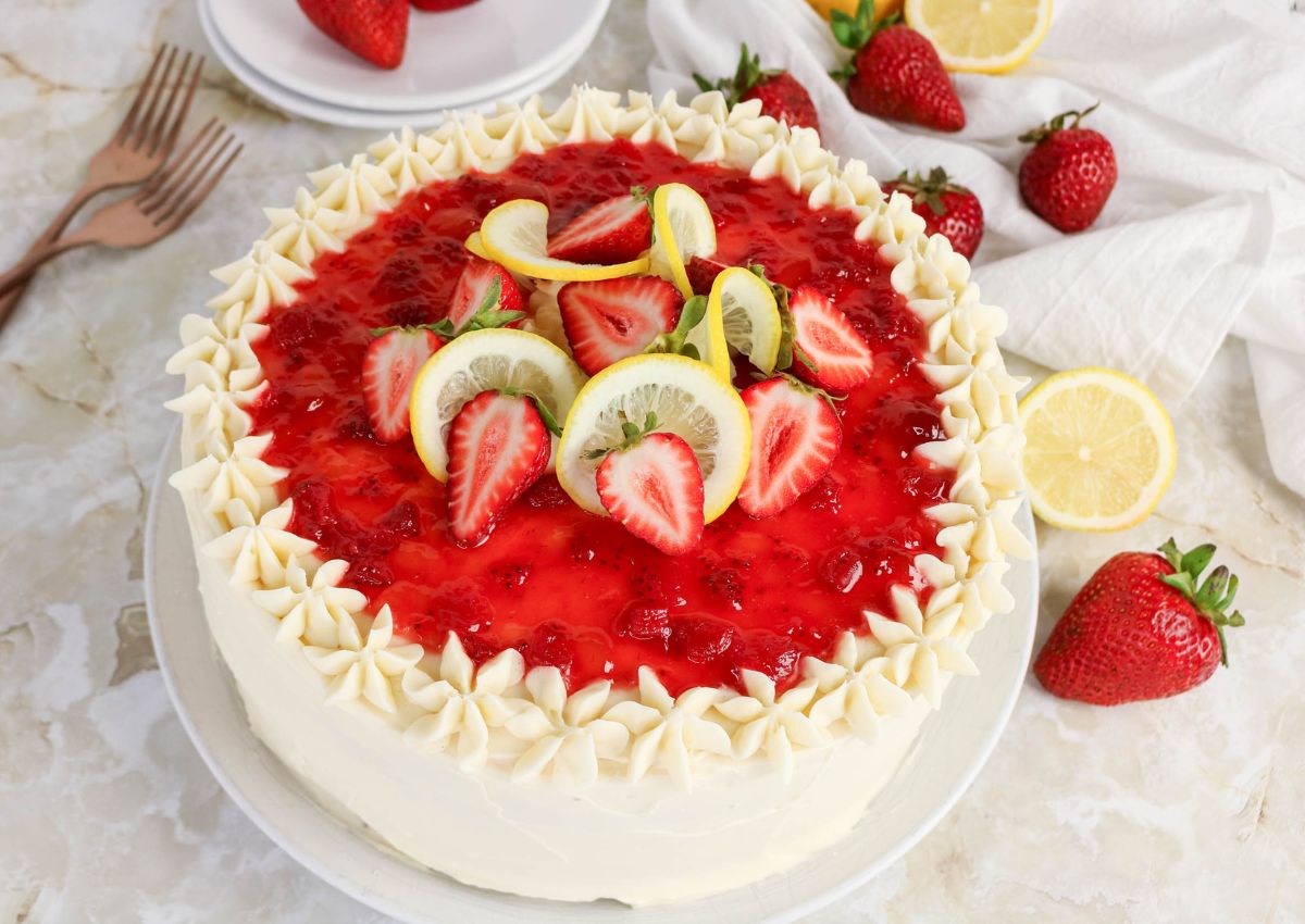 A round cake with white frosting, topped with strawberry glaze, fresh strawberries, and lemon slices, sits on a table surrounded by whole strawberries and lemon slices.