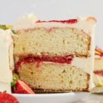 A slice of vanilla layer cake with white frosting and strawberry filling on a white plate, with a fresh strawberry on the side.