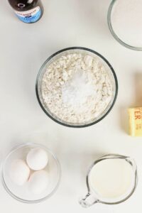 A bowl of flour, eggs, a stick of butter, a measuring cup of milk, a bottle of vanilla extract, and a bowl of sugar arranged on a white surface.