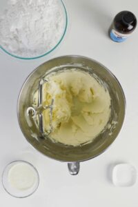 A mixing bowl with creamed butter and a paddle attachment, surrounded by a bowl of powdered sugar, a small bowl of milk, vanilla extract, and an empty dish on a white surface.