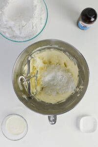 A mixing bowl with butter and powdered sugar, a bowl of flour, a bottle of vanilla extract, a small bowl of milk, and a pinch bowl of salt on a white surface.