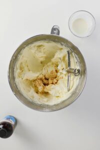 A mixing bowl with whipped butter and vanilla extract, a glass of milk, and a bottle of vanilla extract on a white surface.