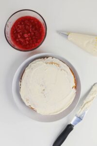 A round cake with white frosting sits on a plate, next to a bowl of strawberry filling, a piping bag, and an icing spatula with frosting.