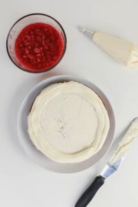A cake with white frosting on a plate, a bowl of red fruit filling, a piping bag, and a spatula with frosting are arranged on a white surface.