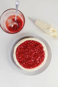 A cake with a layer of red fruit topping and white frosting sits on a plate next to a bowl of fruit mixture with a spoon and a piping bag of frosting.