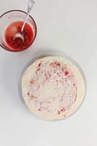 A round cake with white and pink swirled frosting sits next to a glass bowl of mashed strawberries on a white surface.