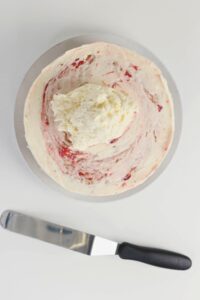 A bowl with partially mixed white and pink frosting sits next to an offset spatula on a white surface.