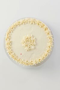 A round cake with smooth white frosting, decorated with piped star-shaped flowers around the edge and center, viewed from above on a white background.
