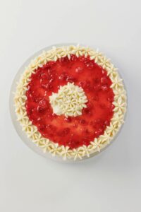 A round cake with a strawberry glaze topping, decorated with white cream rosettes around the edge and in the center, on a white surface.