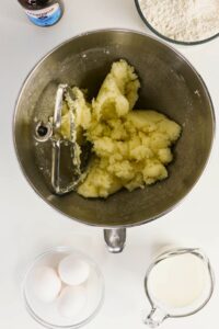 A mixing bowl with creamed butter and sugar, a beater attachment, surrounded by eggs, milk, flour, and vanilla extract on a white surface.