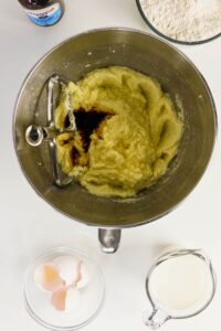 A mixing bowl with creamed sugar and butter, vanilla extract being added, surrounded by a bowl of flour, a jug of milk, and broken eggs in a bowl.