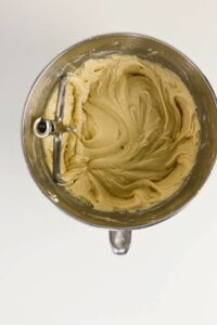 A metal mixing bowl with creamy batter and a paddle attachment, viewed from above on a white surface.