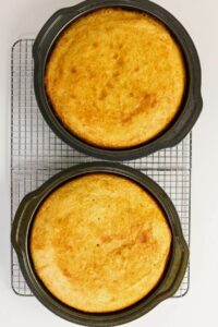 Two round golden-brown cakes in baking pans cooling on a metal wire rack against a white background.