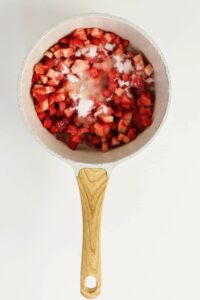 A saucepan with chopped strawberries and sugar on top, placed on a white background, viewed from above.