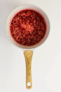 A saucepan with a wooden handle containing chopped strawberries simmering in liquid on a white background.