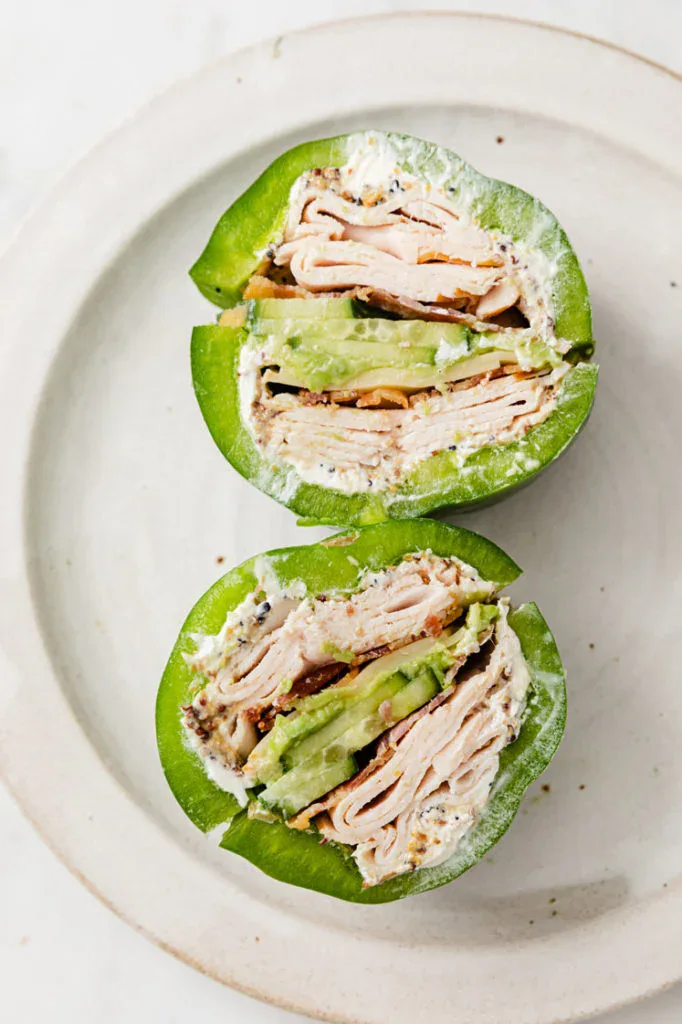 Halved green bell pepper stuffed with sliced deli turkey, avocado, cream cheese, and herbs, arranged on a white plate.