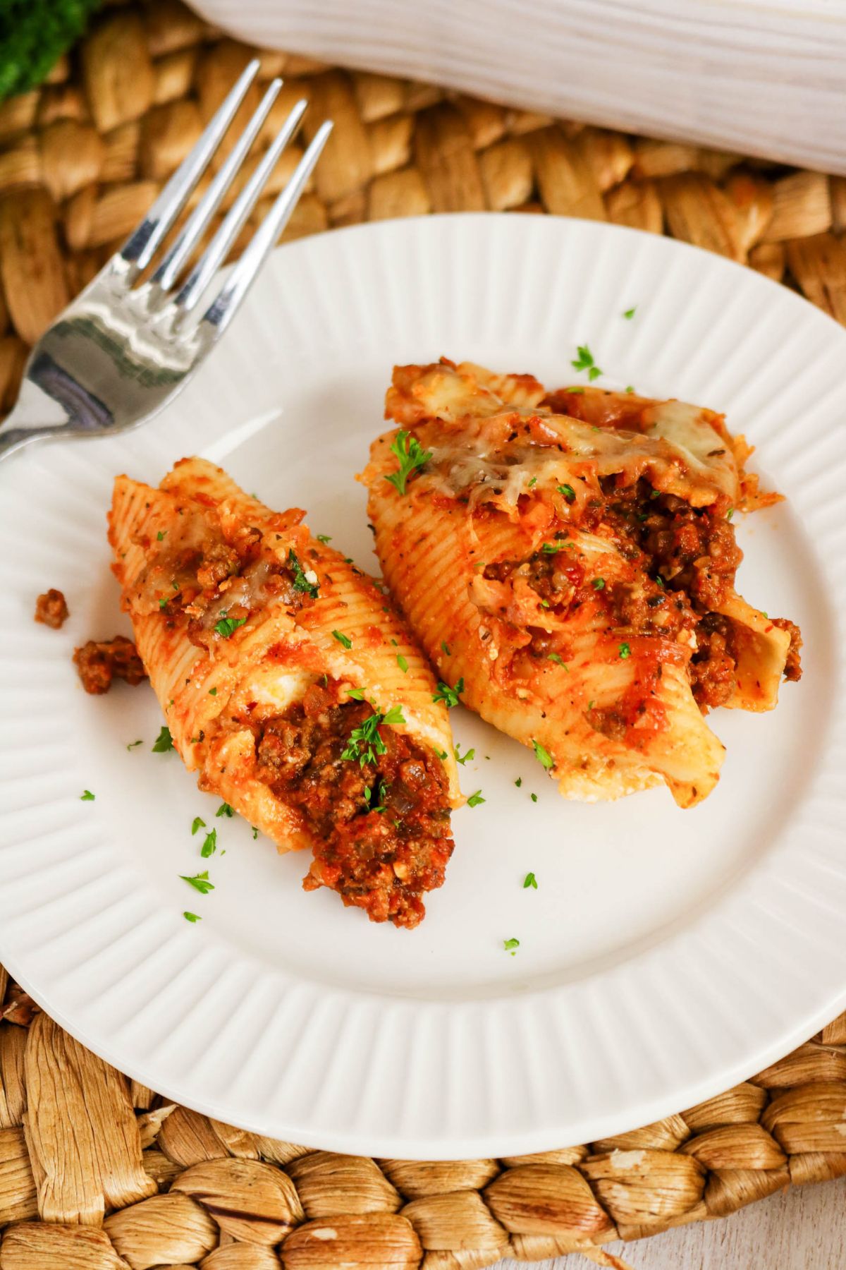 Two stuffed pasta shells with meat sauce and cheese on a white plate, garnished with chopped herbs, next to a fork on a woven placemat.