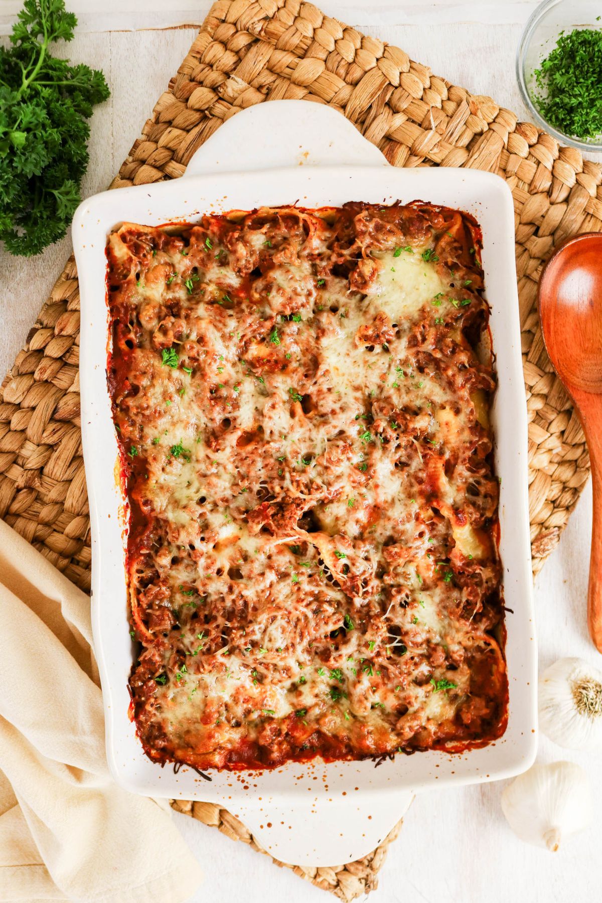 A baked lasagna in a white rectangular dish on a woven mat, topped with melted cheese and herbs, with a wooden spoon and fresh parsley nearby.