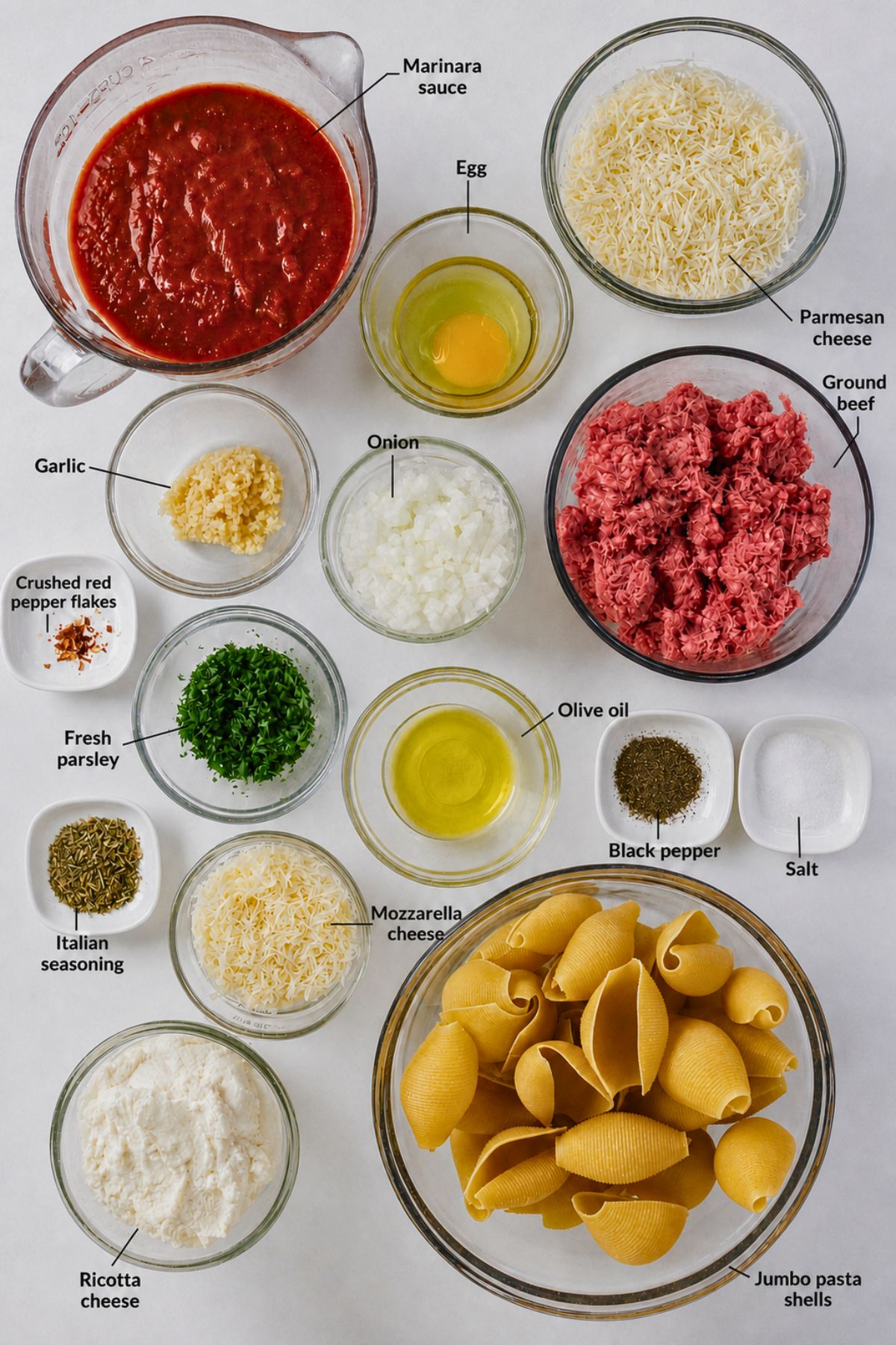 Ingredients for stuffed shells arranged on a white surface, including marinara sauce, egg, cheeses, ground beef, onion, garlic, parsley, olive oil, seasonings, and jumbo pasta shells.