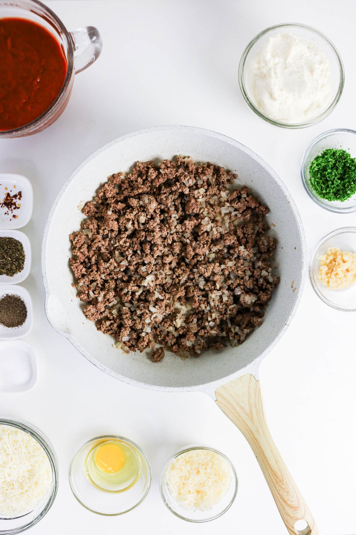 Cooked ground beef in a white skillet surrounded by bowls of tomato sauce, ricotta cheese, herbs, shredded cheese, an egg, spices, and minced garlic on a white surface.