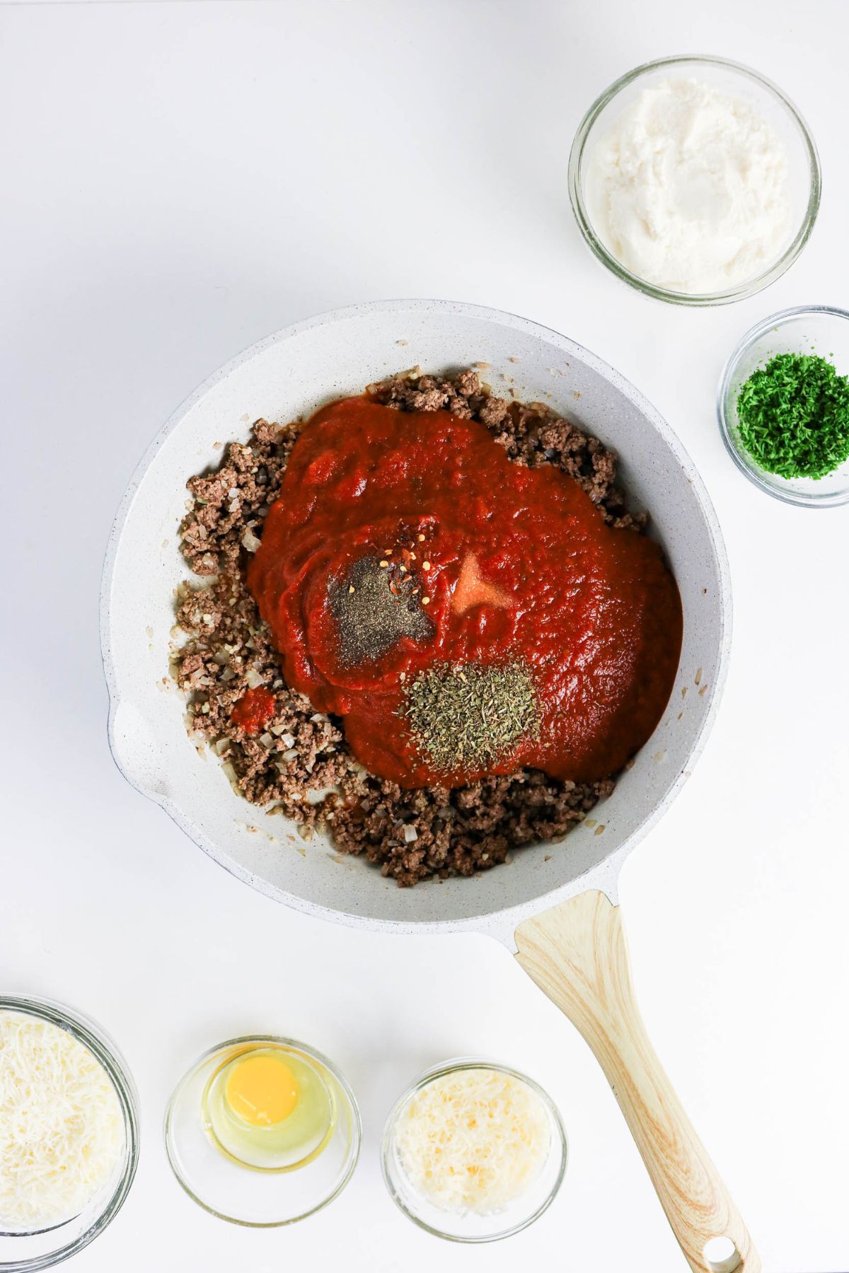 A pan with cooked ground beef topped with tomato sauce and herbs, surrounded by bowls of ricotta, parmesan, an egg, chopped parsley, and shredded cheese on a white surface.