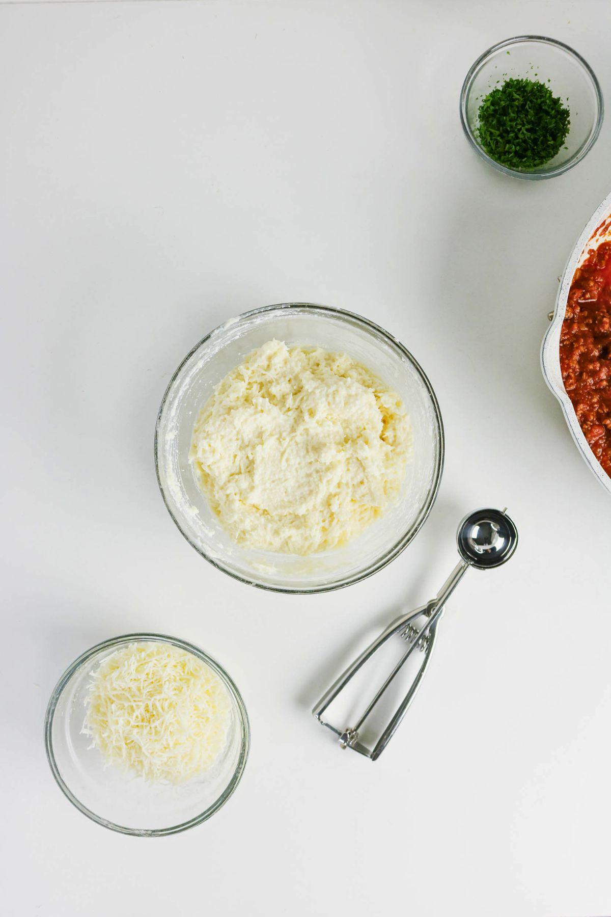 A glass bowl of cheese mixture, a bowl of shredded cheese, a bowl of chopped herbs, a metal scoop, and a dish of red sauce on a white surface.