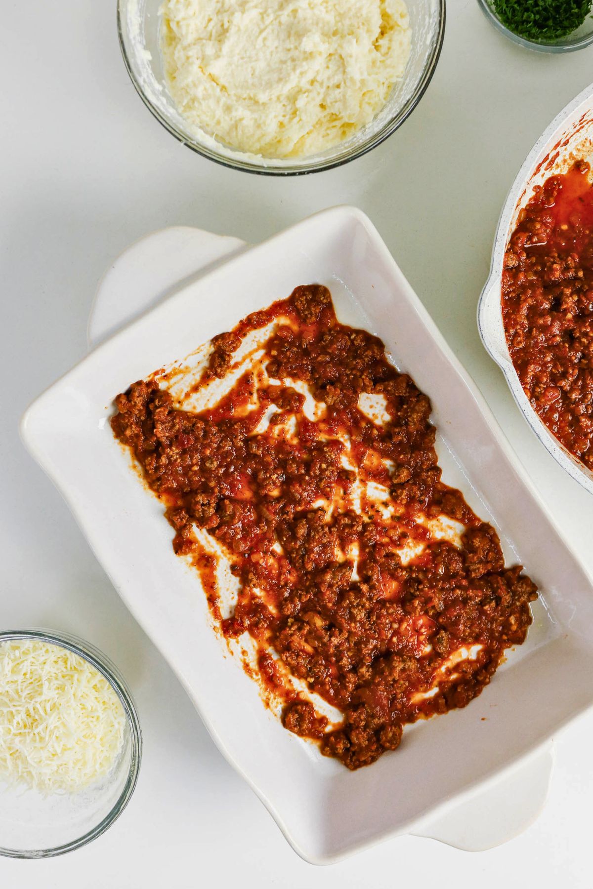 A baking dish with a layer of meat sauce, surrounded by bowls of shredded cheese, ricotta mixture, and chopped herbs on a white surface.