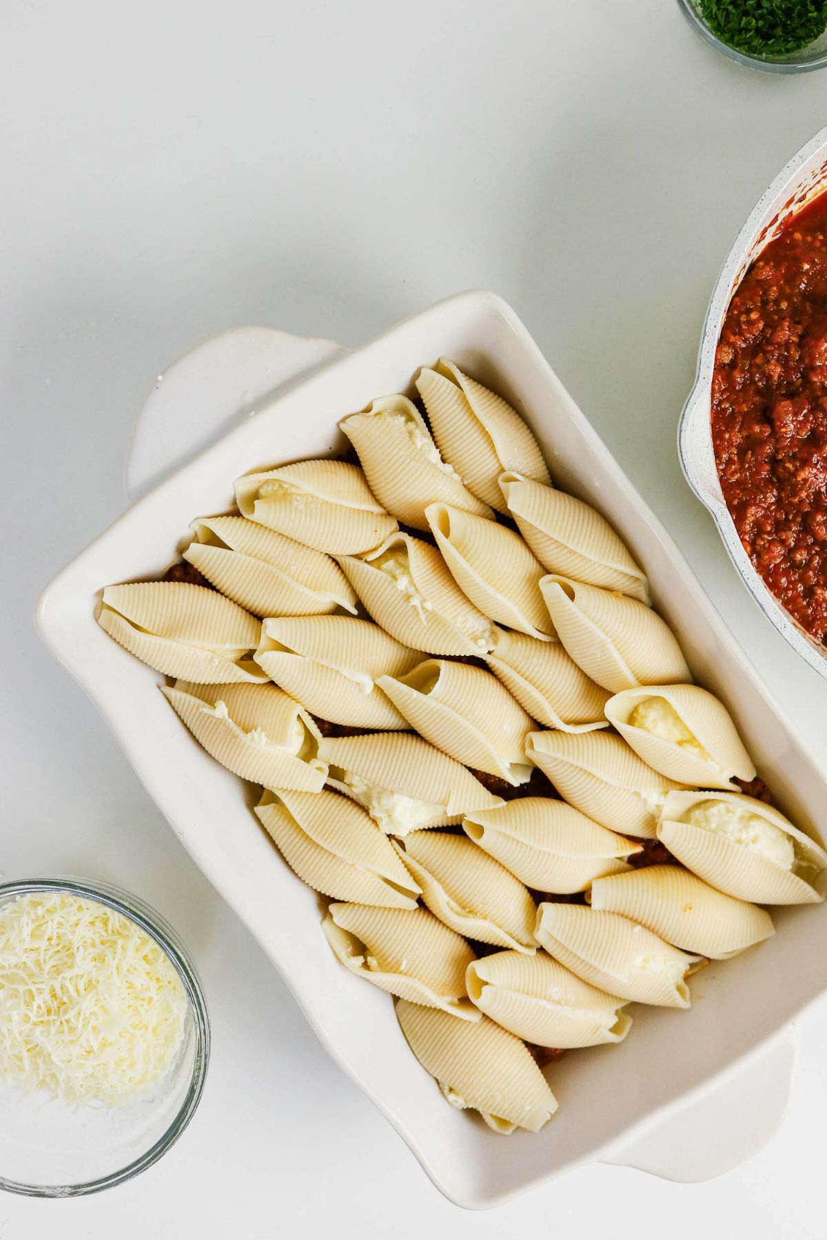 Uncooked pasta shells filled with cheese arranged in a white baking dish, next to a bowl of shredded cheese and a pot of red meat sauce.