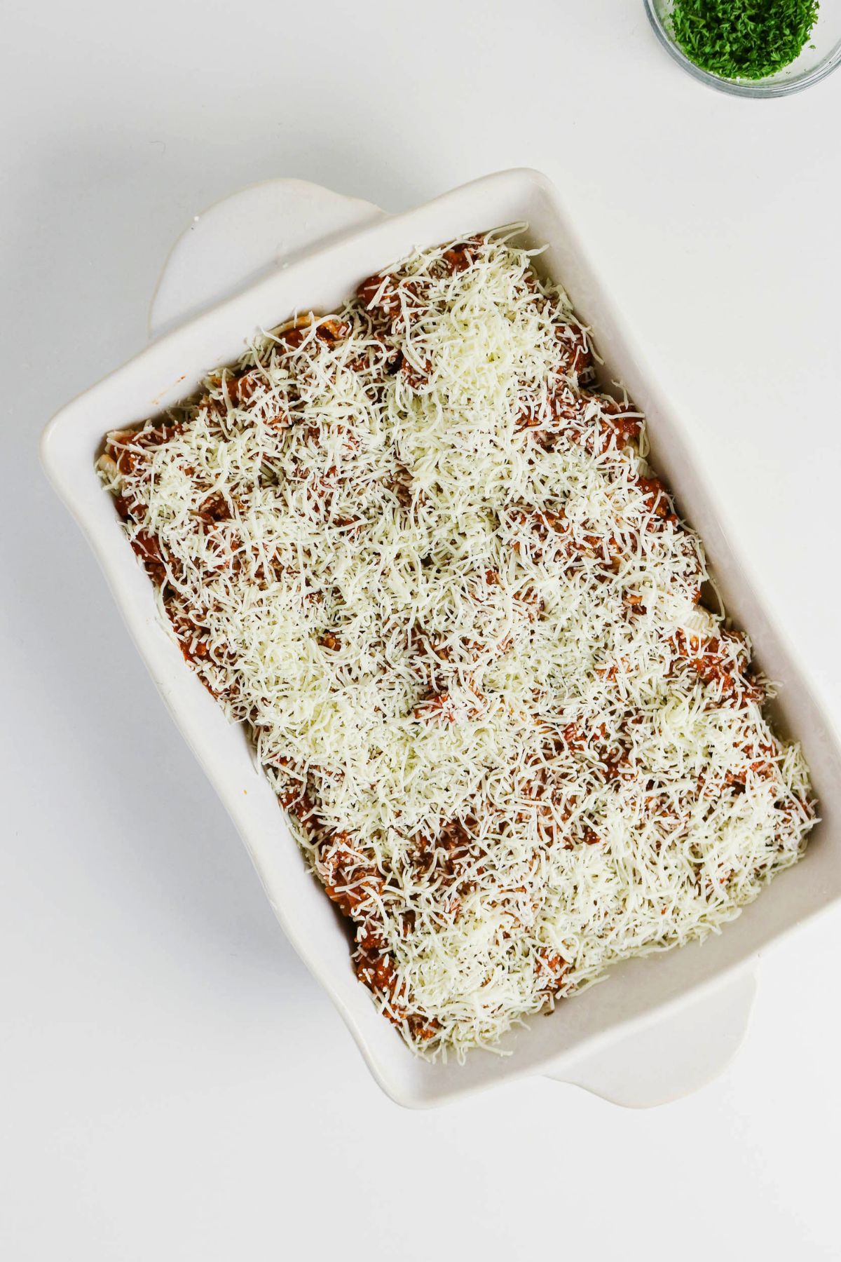 A rectangular white baking dish filled with food topped with shredded cheese, placed on a white surface.