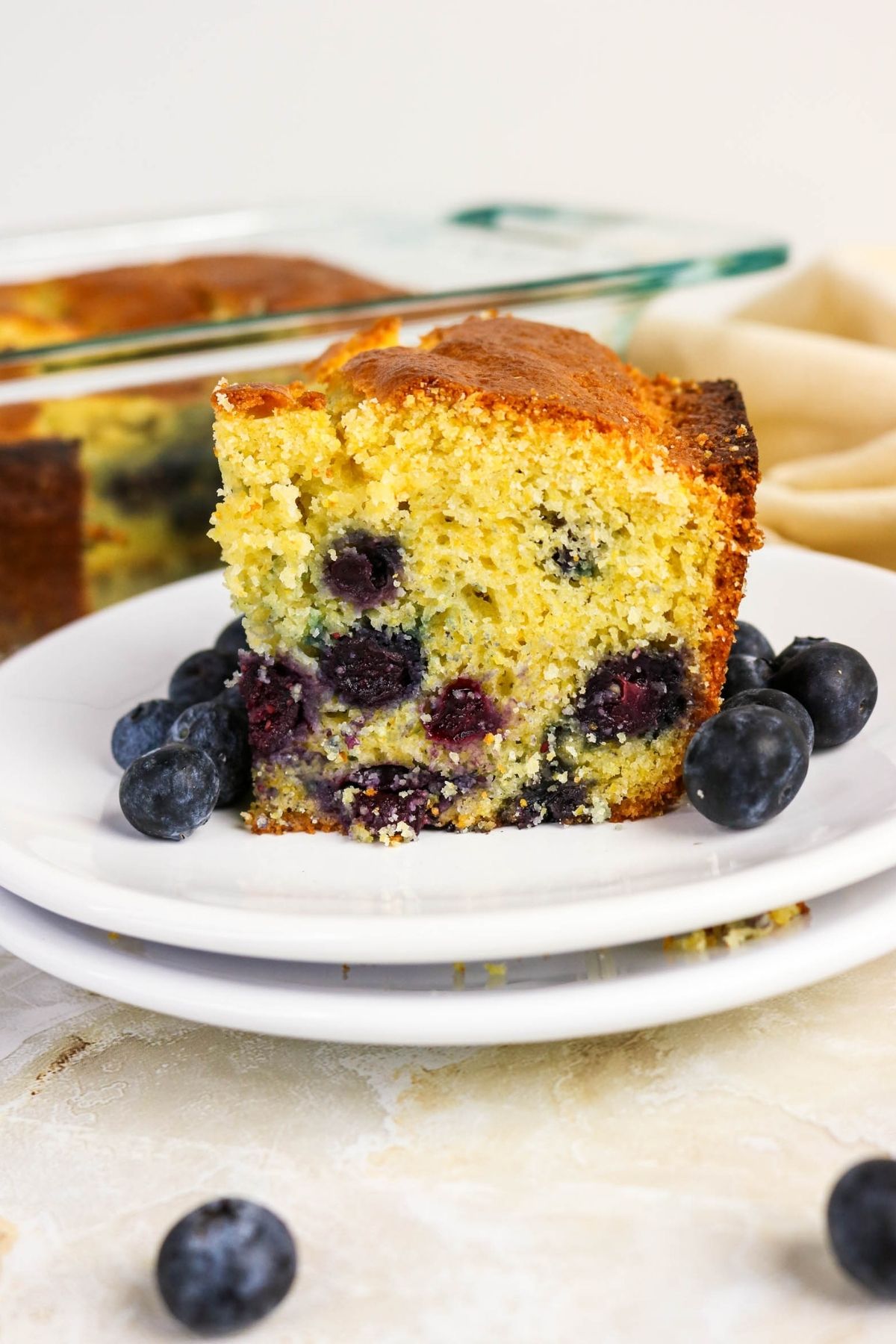 A slice of blueberry cornbread on stacked white plates, with fresh blueberries scattered around.