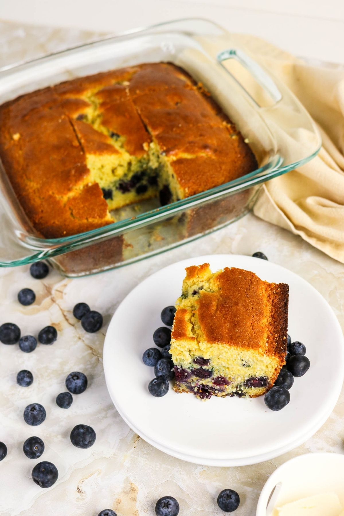 A square piece of blueberry cake on a white plate with fresh blueberries, next to a glass baking dish containing the rest of the cake.