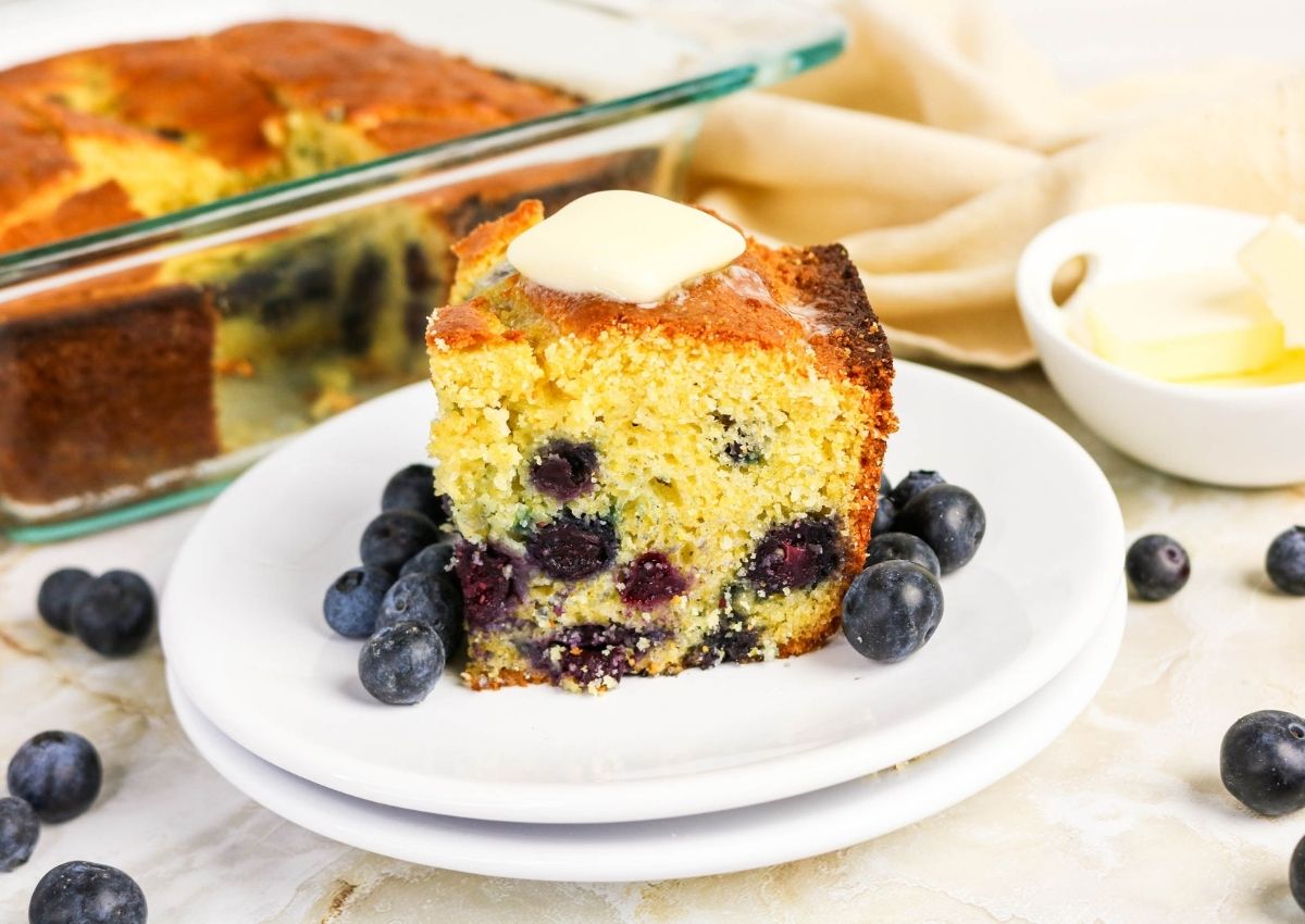 A slice of blueberry cornbread with a pat of butter on top, served on a white plate with fresh blueberries, next to a baking dish and a bowl of butter.