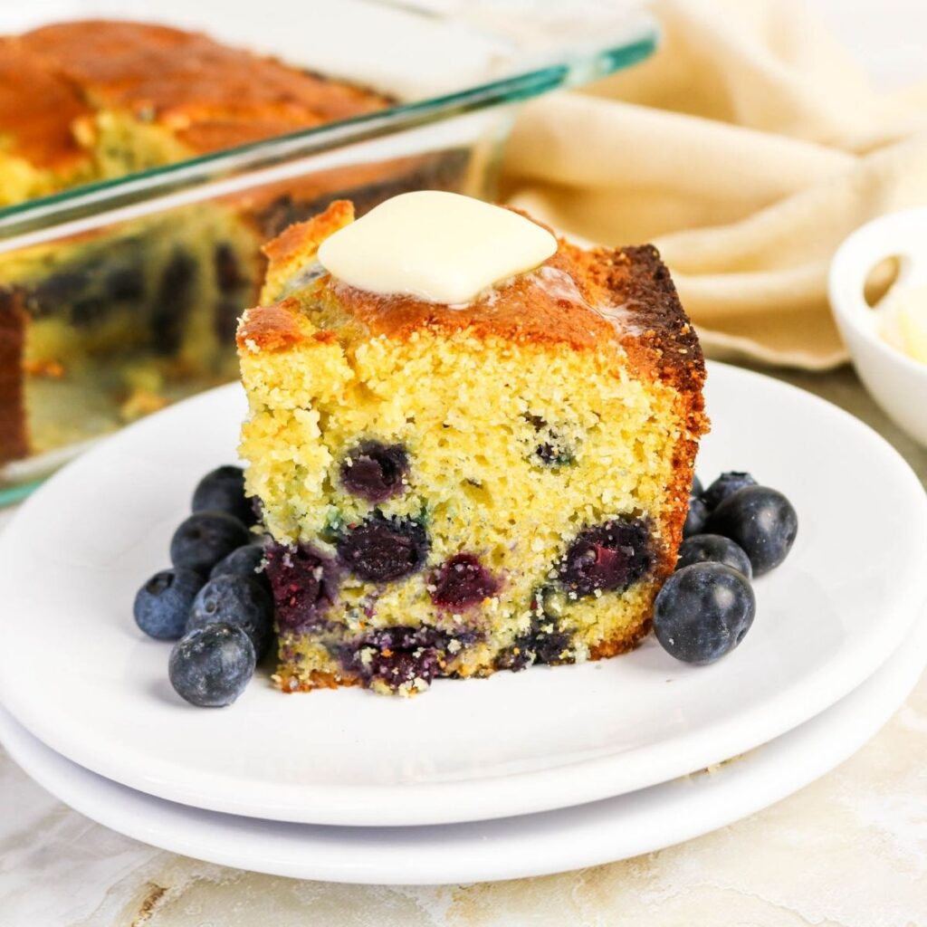 A slice of blueberry cornbread with melted butter on top, served on a white plate with fresh blueberries.