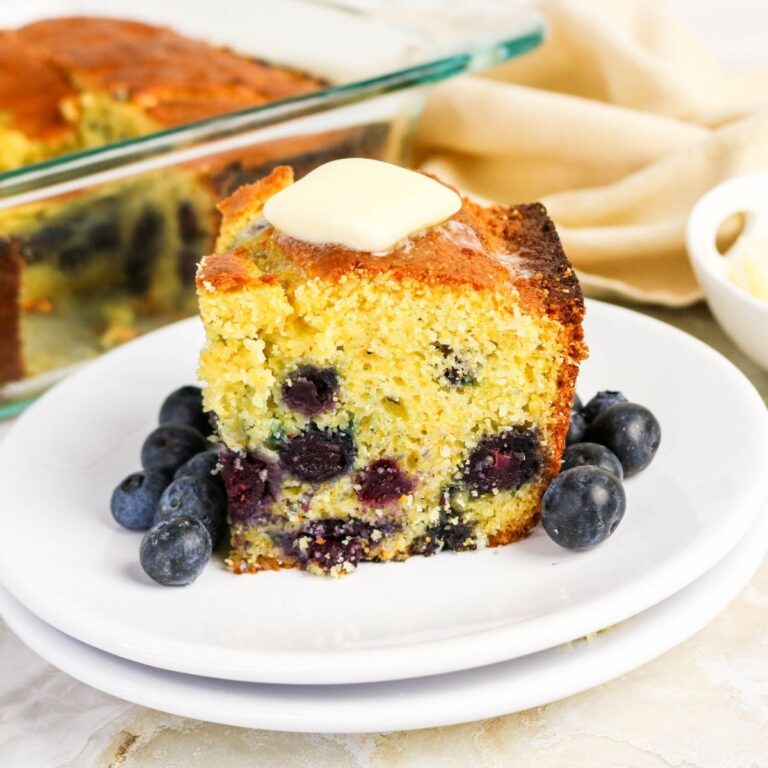 A slice of blueberry cornbread with melted butter on top, served on a white plate with fresh blueberries.