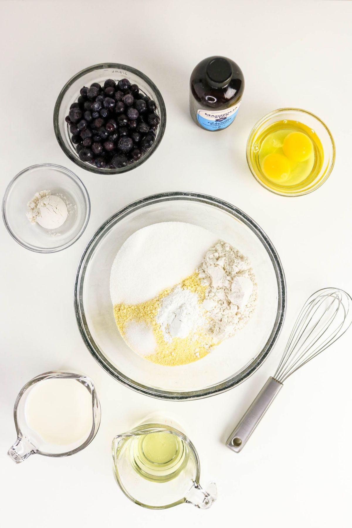 Bowls of flour, sugar, cornmeal, baking powder, eggs, blueberries, a bottle of vanilla extract, milk, oil, and a whisk are arranged on a white surface.