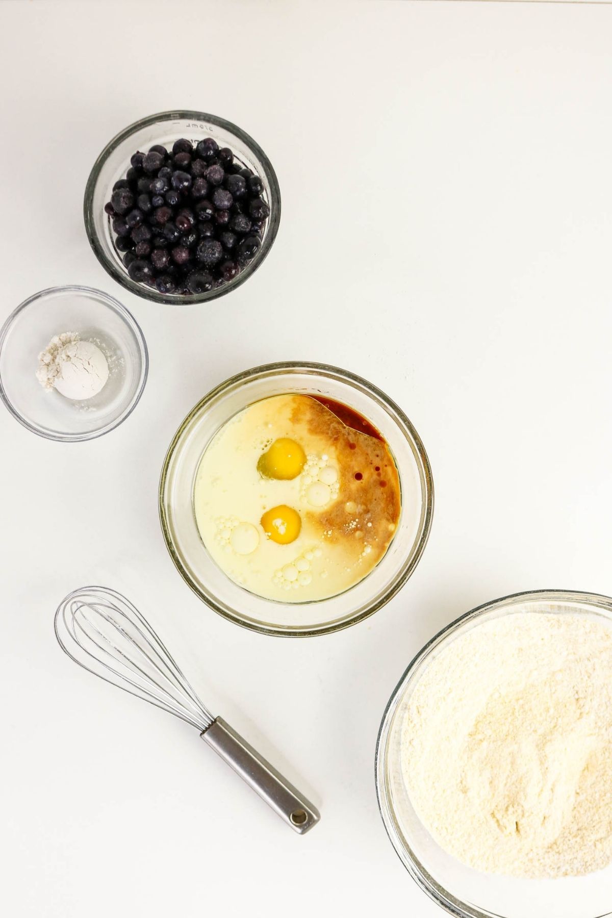Bowl with eggs, milk, and vanilla, bowl of flour, bowl of blueberries, small bowl of baking powder, and a whisk on a white surface.
