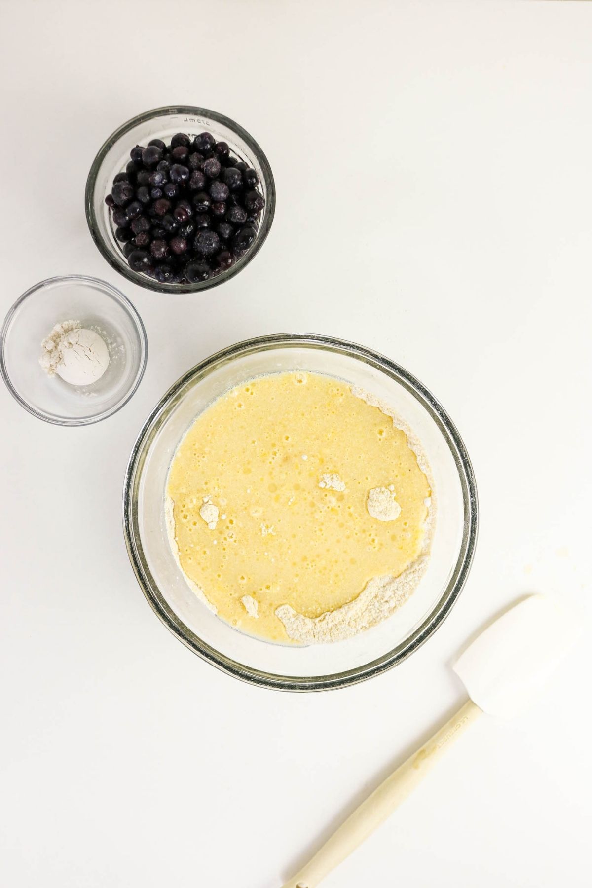 A bowl of pancake batter with some flour on top, a bowl of blueberries, a small bowl of flour, and a spatula on a white surface.