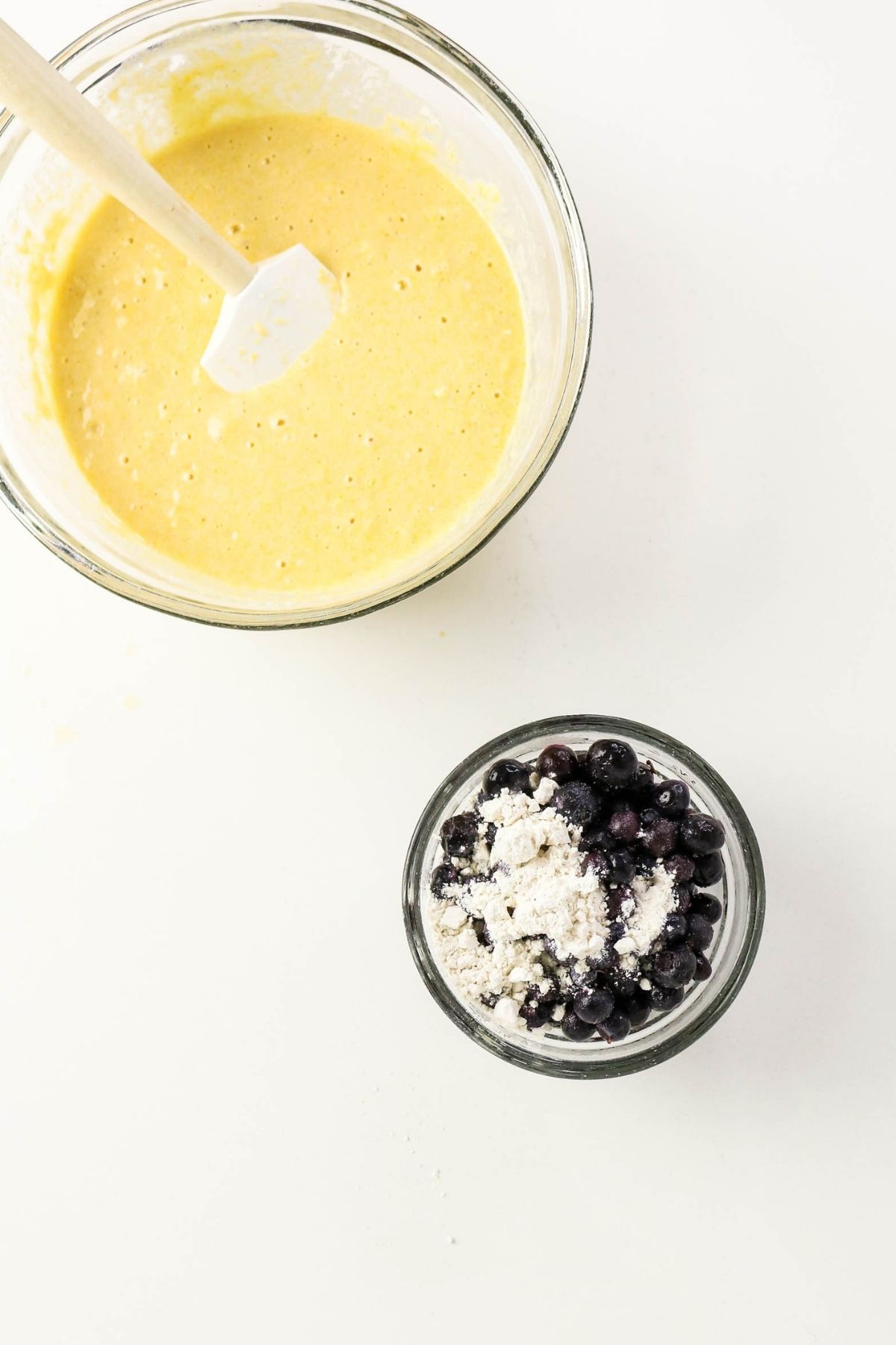 A bowl of yellow batter with a spatula next to a smaller bowl containing blueberries mixed with flour, on a white surface.