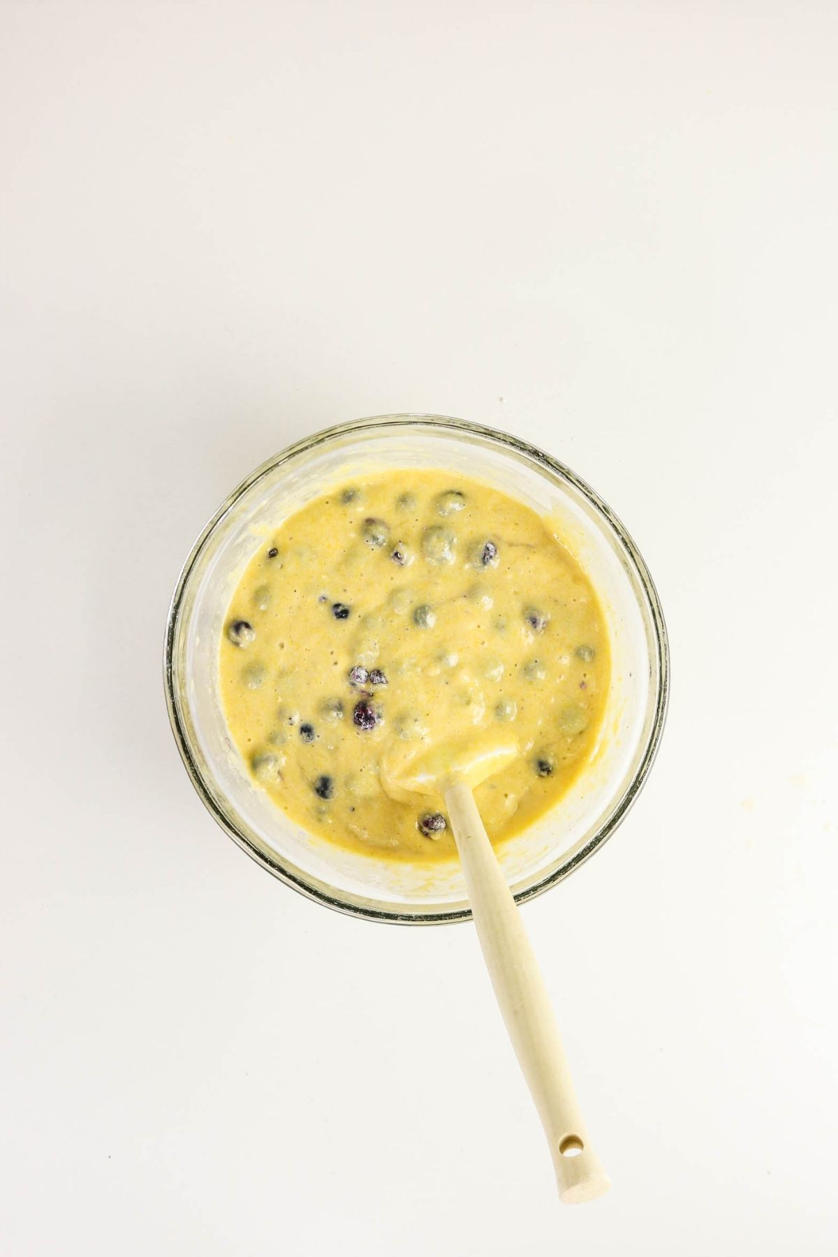 A glass bowl filled with blueberry muffin batter and a white spoon, viewed from above on a white background.