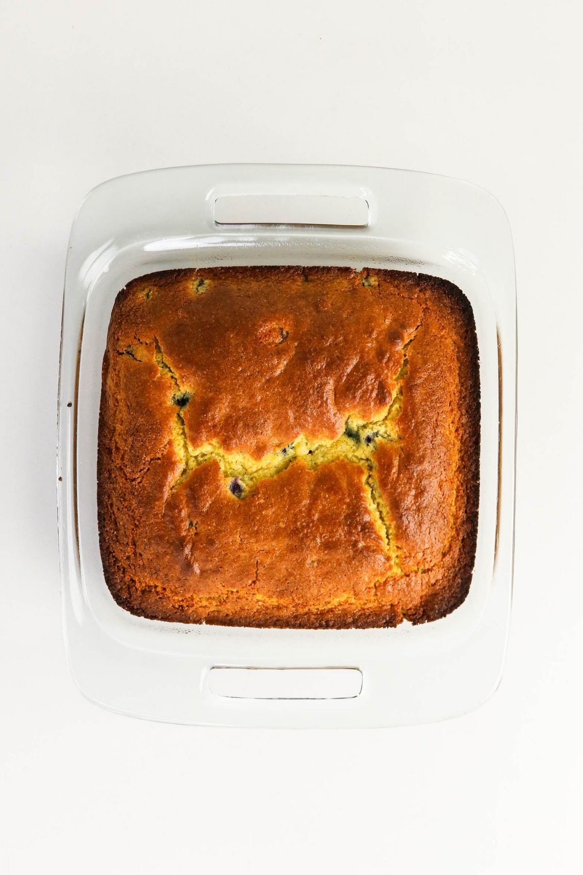 A golden-brown rectangular loaf cake with cracks on top, baked in a white ceramic dish, shown from above on a white background.