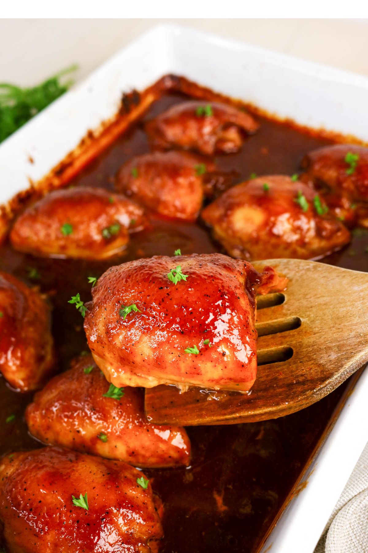 Baked chicken thighs coated in a glossy barbecue sauce, garnished with chopped herbs, in a white baking dish with a wooden spatula lifting one piece.