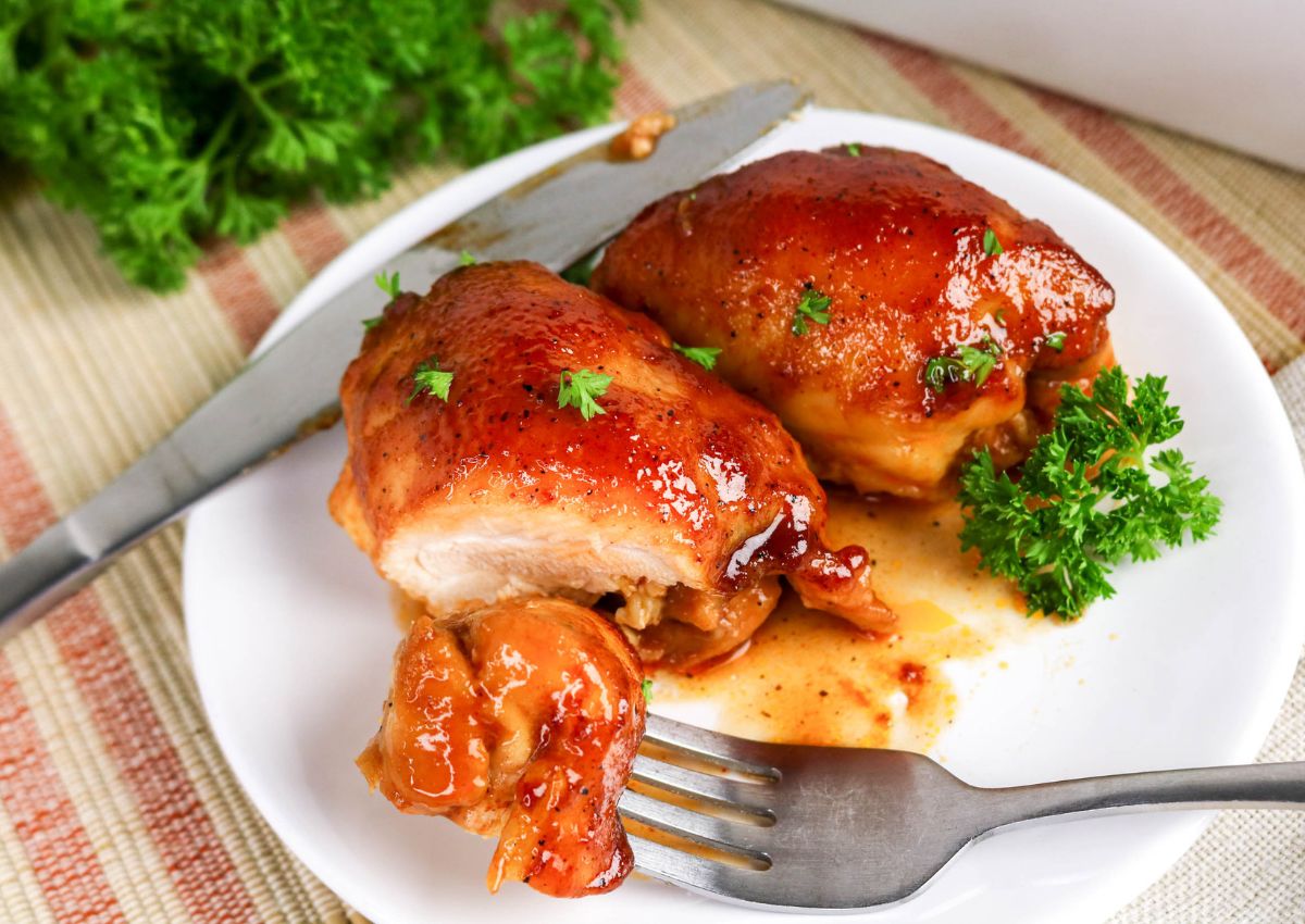 Two glazed, cooked chicken thighs on a white plate with sauce, garnished with parsley. A knife and fork are beside the chicken.