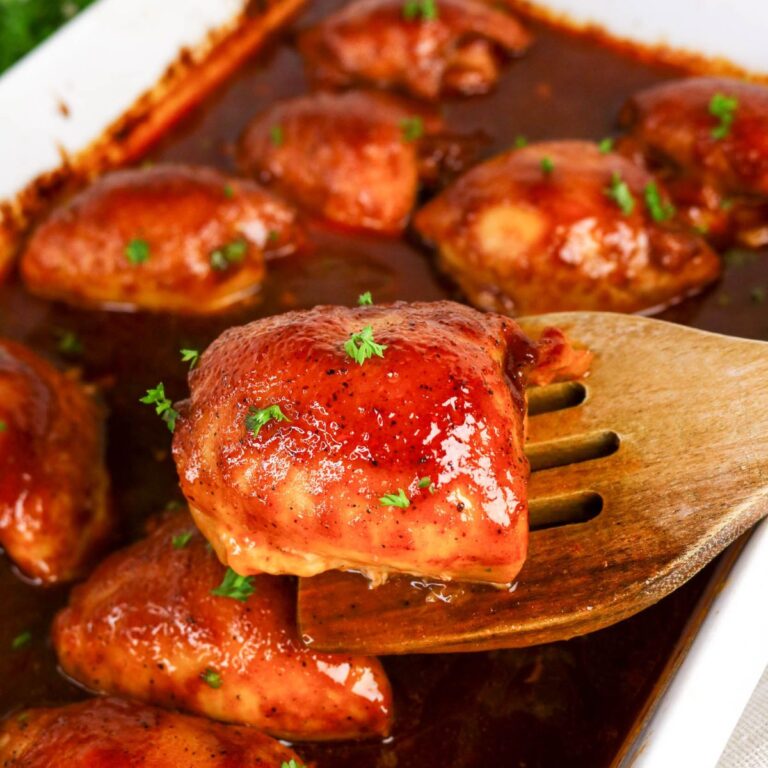 Baked chicken thighs coated in a reddish-brown sauce, garnished with chopped herbs, with one piece being lifted by a wooden spatula from a white baking dish.