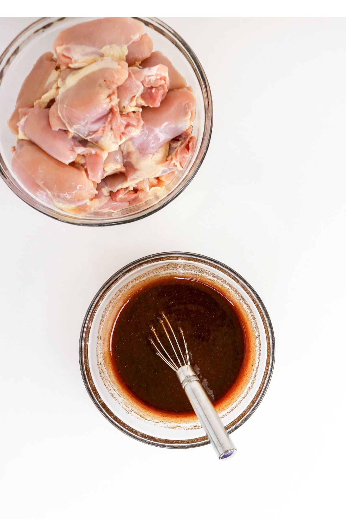 Two glass bowls: one with raw chicken thighs, the other with a dark marinade and a whisk, on a white surface.