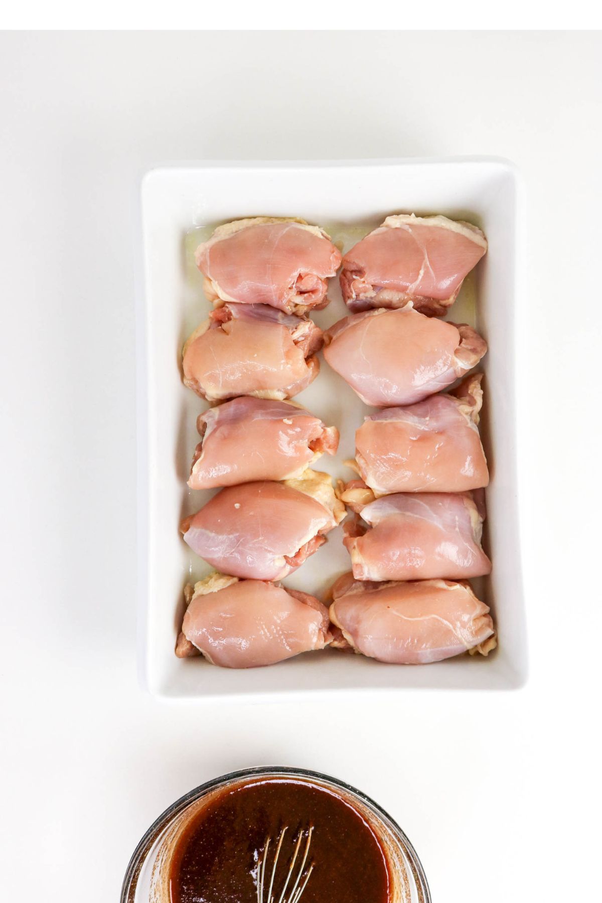 A rectangular white dish holds ten raw boneless chicken thighs, arranged in two rows; a bowl of brown marinade with a whisk is placed below the dish.