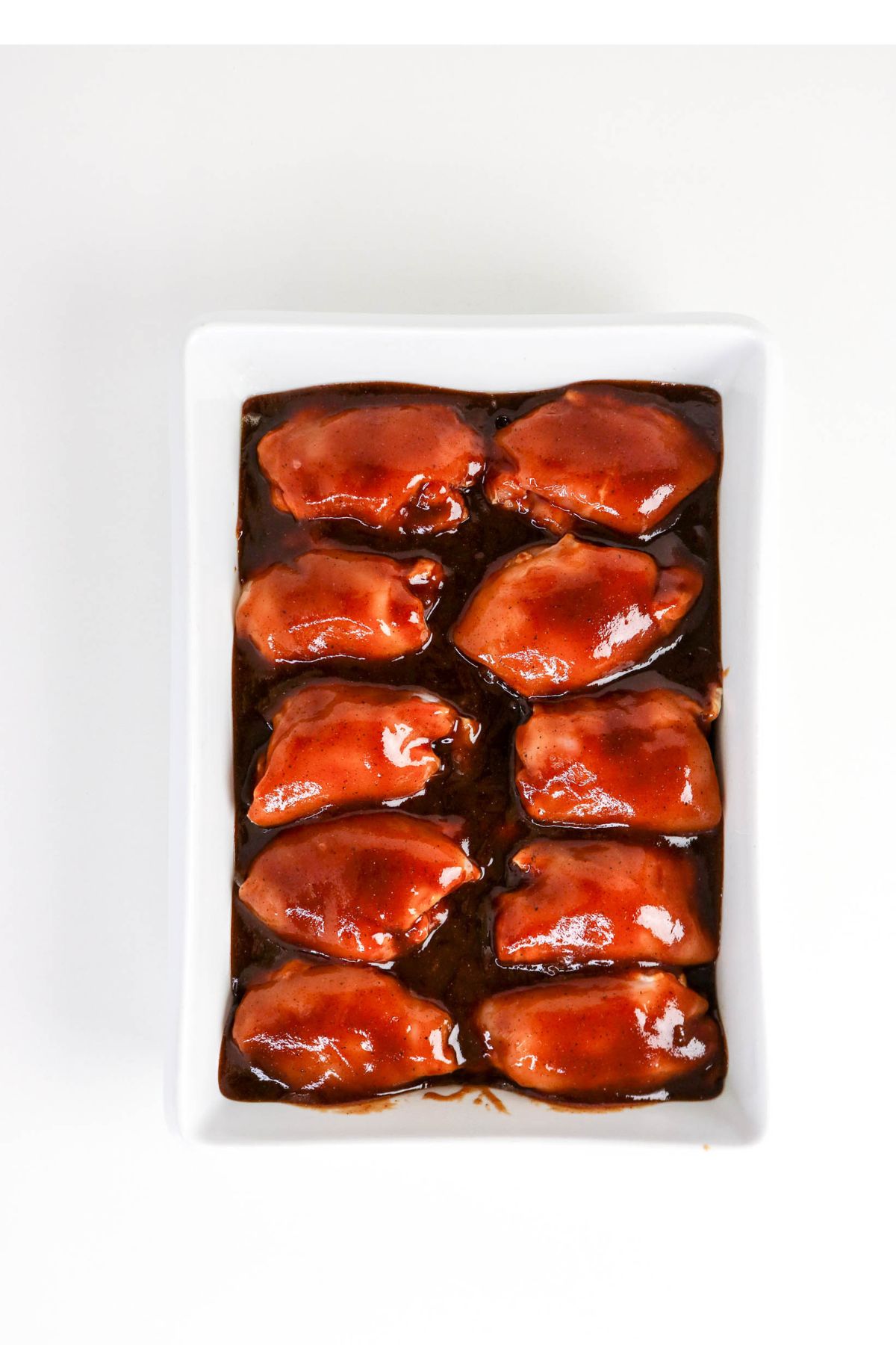Rectangular white dish with ten pieces of glazed chicken thighs arranged in two rows, coated in a dark sauce, on a white background.